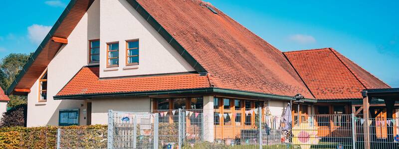 Die Ortsverwaltung und der Kindergarten in Bussmannshausen mit rotem Ziegeldach steht hinter einem Zaun und einer niedrigen Steinmauer, umgeben von blühenden Pflanzen, unter einem klaren blauen Himmel.