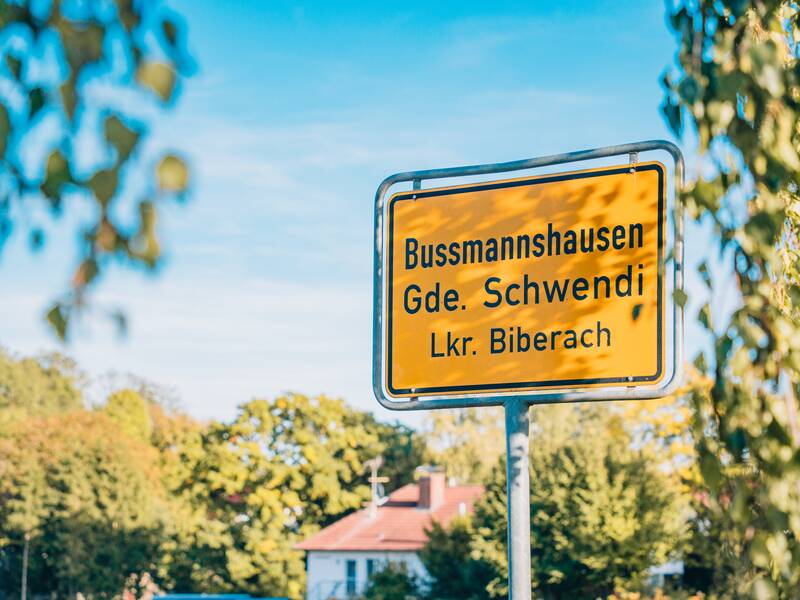 Ein gelbes Ortsschild zeigt die Ortschaft "Bussmannshausen" in der Gemeinde Schwendi im Landkreis Biberach, umgeben von Bäumen und blauem Himmel.