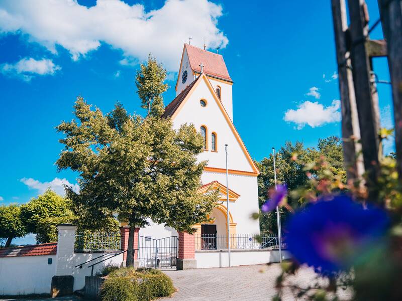 Eine kleine, weiße Kirche mit einem roten Dach steht umgeben von Bäumen unter einem strahlend blauen Himmel, im Vordergrund sind unscharfe bunte Blumen zu sehen.