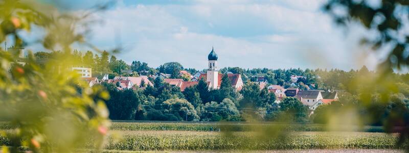 Schwendi mit einer Kirche im Zentrum ist von grünen Feldern und Bäumen umgeben, während der Himmel von Wolken bedeckt ist.