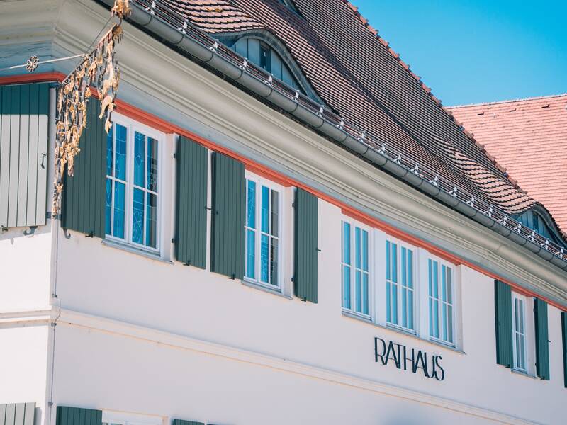 Das Bild zeigt den oberen Teil eines Gebäudes mit der Aufschrift "Rathaus", grünen Fensterläden und einem roten Ziegeldach unter klarem blauen Himmel.