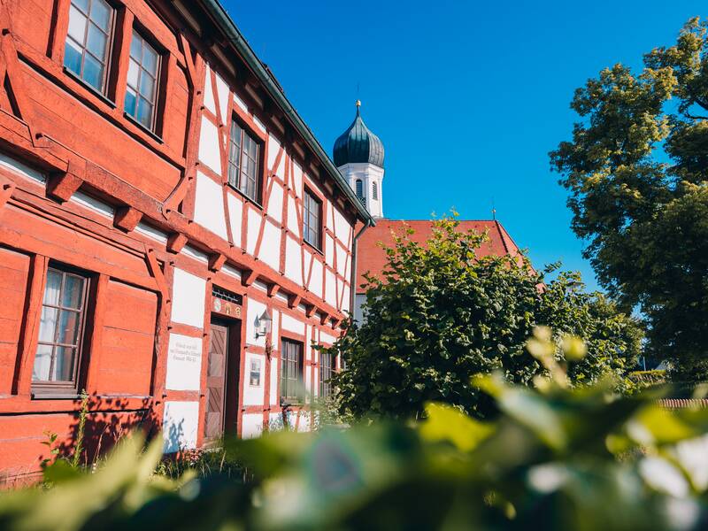 Das Pfarrhaus, ein rot-weißes Fachwerkhaus, steht neben einer Kirche mit Zwiebelturm, umgeben von grünen Bäumen und Sträuchern unter einem klaren blauen Himmel.