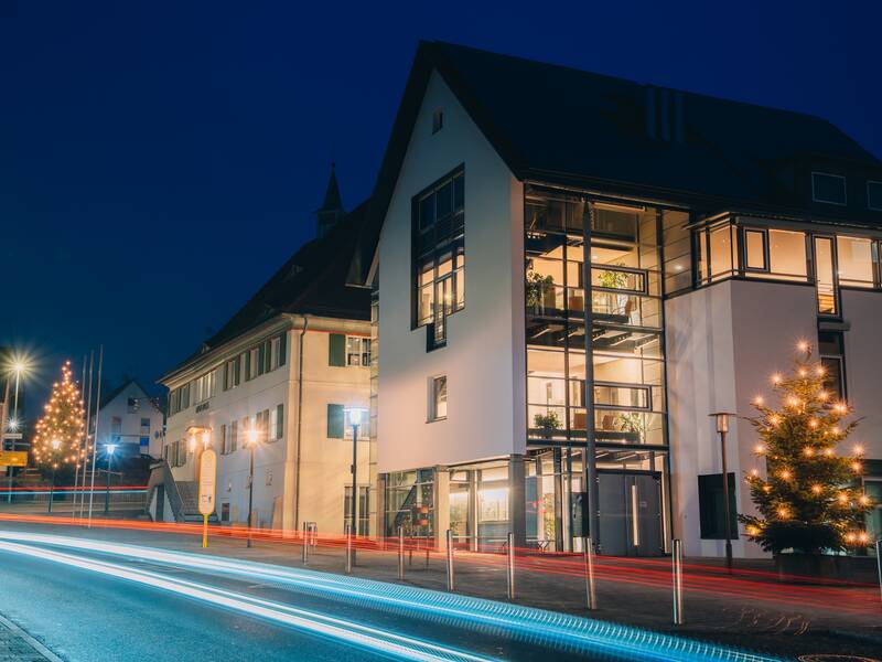 Ein modernes Gebäude ist an einem Abend festlich mit Weihnachtslichtern geschmückt und von durchgehenden Lichtspuren vorbeifahrender Fahrzeuge umgeben.