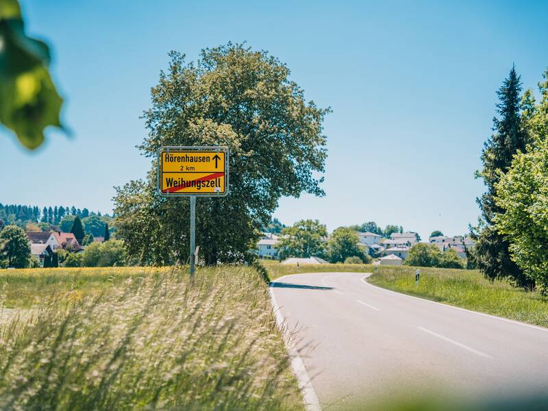 Eine Landstraße führt durch eine grüne Landschaft mit einem gelben Schild, das Entfernungen zu nahegelegenen Orten anzeigt.